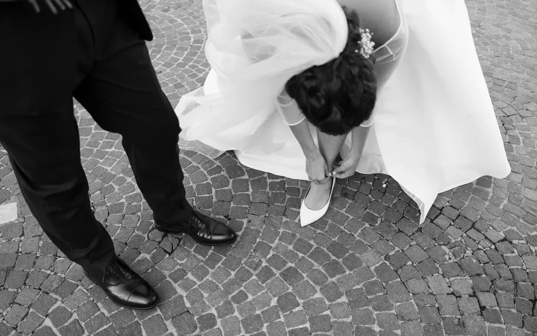Sposa in abito bianco mentre si allaccia la scarpa, con lo sposo accanto, su una pavimentazione in pietra